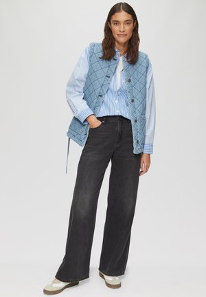 Vrouw draagt een blauw doorgestikt vest over een blauw gestreept shirt met zwarte wijde jeans en witte sneakers, staand met één hand in de zak.