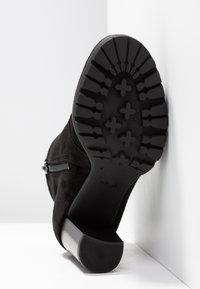 Botte noire à talon inclinée contre un mur, montrant une semelle en caoutchouc épaisse avec un profil profond et une fermeture éclair latérale.