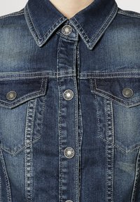 Blå denimjacka med en klassisk krage, två bröstfickor med klaffar och knappstängning med metalliska detaljer. Faded texturdetalj.