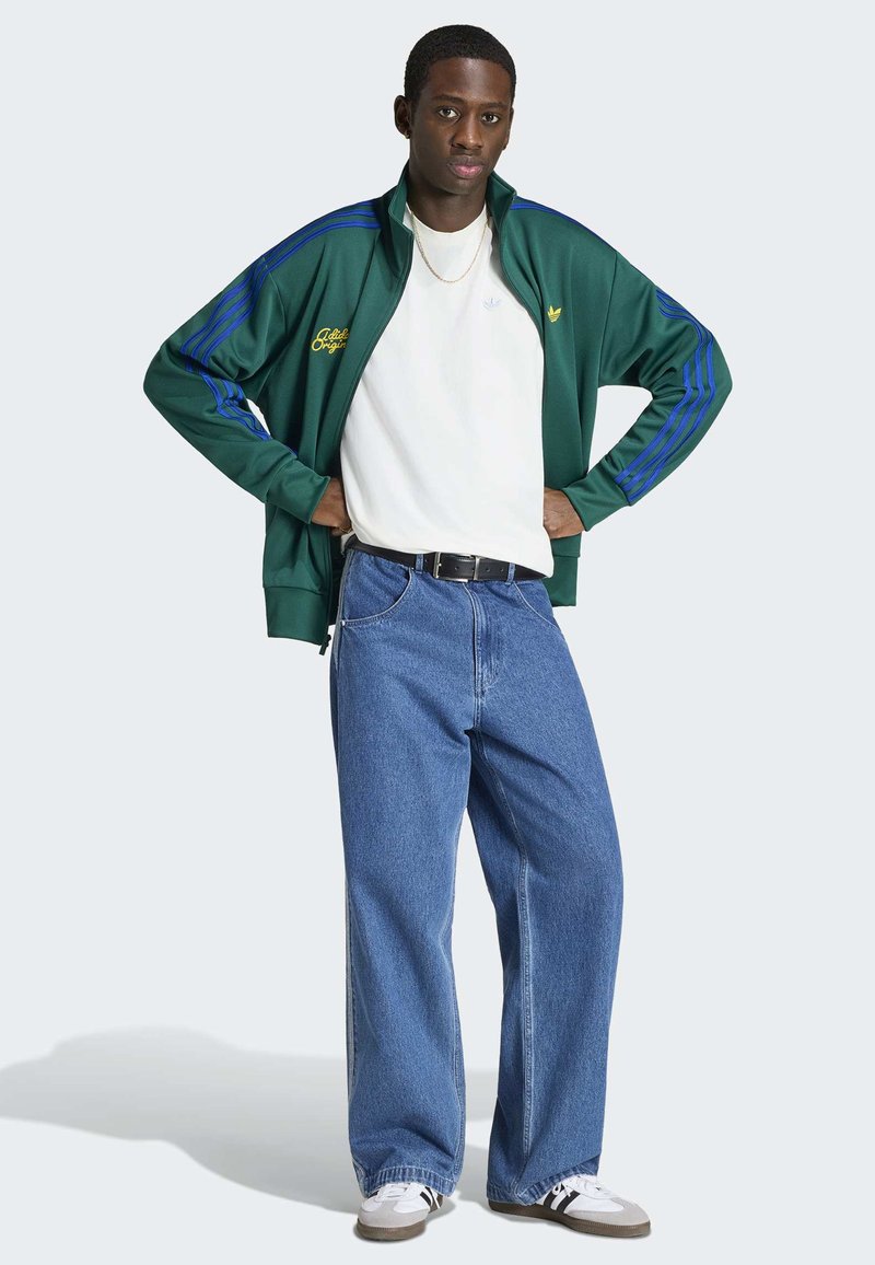 Mann steht mit Händen in den Hüften, trägt eine grüne und blaue Trainingsjacke, weißes T-Shirt, lockere blaue Jeans und weiße Turnschuhe vor grauem Hintergrund.