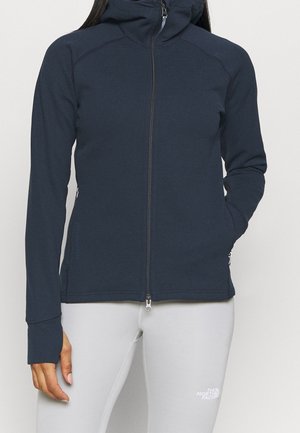Persoon met een marineblauwe hoodie met rits en capuchon en lichtgrijze broek, linkerhand zichtbaar, staand tegen een effen lichte achtergrond.