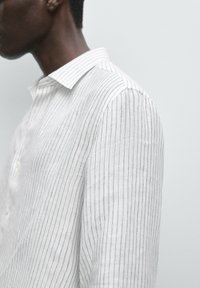 Vue de côté d'une personne portant une chemise blanche à manches longues avec de fines rayures noires verticales, sur un fond clair uni.