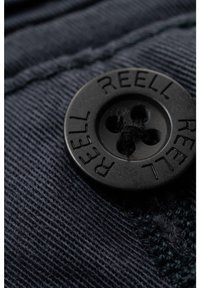 Botón negro de cuatro agujeros con la etiqueta "REELL" cosido sobre tela texturizada azul oscuro, primer plano.