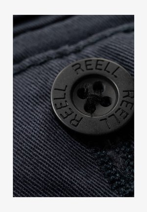 Black plastic button with four holes, engraved with the word "REELL," positioned on textured dark fabric with visible stitching.
