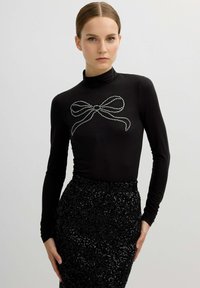 Top negro de manga larga con cuello perkins, adornado con un diseño de lazo de pedrería plateada en el pecho, combinado con una falda negra de lentejuelas.