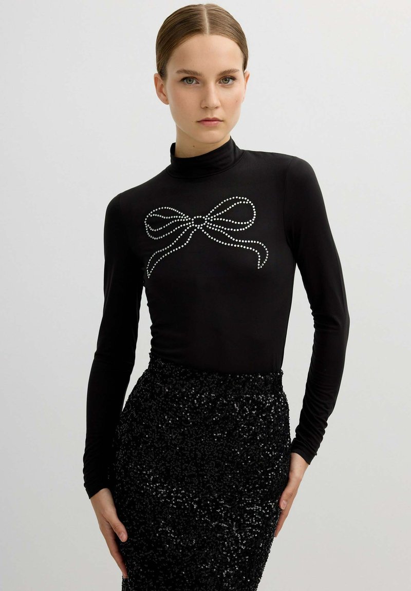 Top negro de manga larga con cuello perkins, adornado con un diseño de lazo de pedrería plateada en el pecho, combinado con una falda negra de lentejuelas.