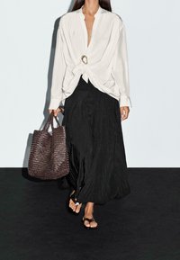 Chemise blanche à manches longues avec un devant noué et un accent doré, associée à une jupe maxi plissée noire, portant un sac cabas tissé marron foncé.