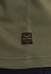 Una tela texturizada de color verde oliva presenta una etiqueta rectangular negra con letras doradas que dice "PME LEGEND" y "COMPROBADO A GRAN ALTITUD".