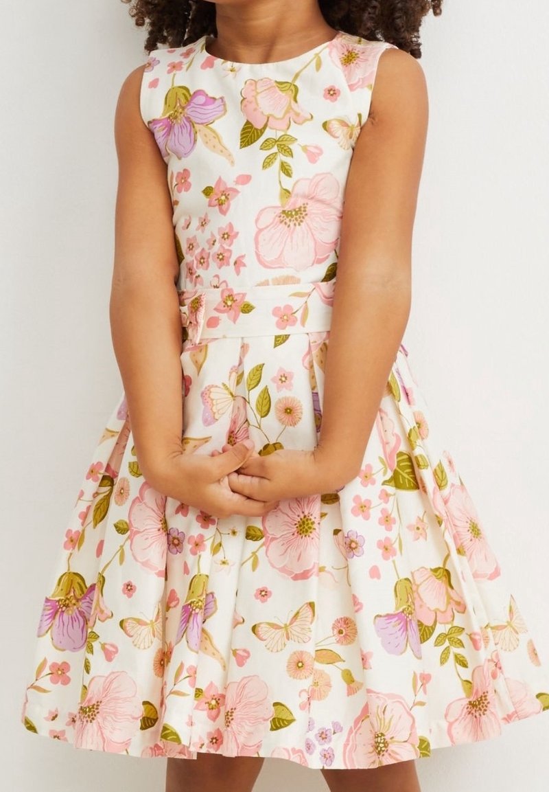 Jeune fille portant une robe blanche sans manches avec des motifs floraux et papillons roses, les mains jointes devant.