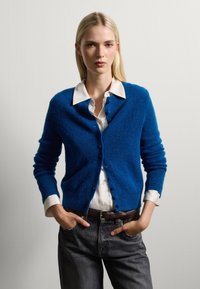 Blauwe gebreide cardigan met gestructureerde stof, ronde halslijn en knoopsluiting aan de voorkant, gedragen over een witte blouse met kraag en grijze jeans.