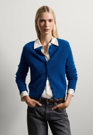 Cardigan blu lavorato a maglia con tessuto testurizzato, scollatura rotonda e chiusura con bottoni, indossato sopra una camicia bianca con colletto e jeans grigi.