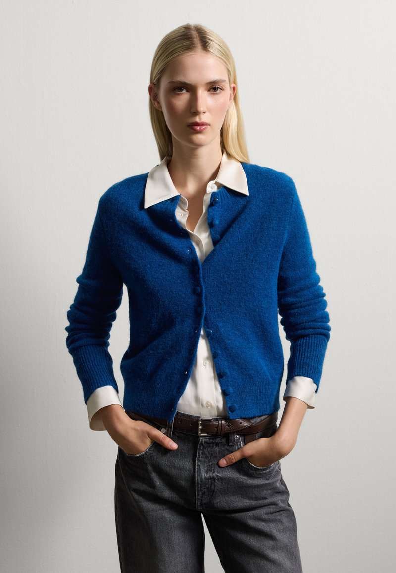 Blauwe gebreide cardigan met gestructureerde stof, ronde halslijn en knoopsluiting aan de voorkant, gedragen over een witte blouse met kraag en grijze jeans.