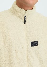 Beige Fleece-zipperjacke mit hohem Kragen, ausgestattet mit einem schwarzen Markenlabel. Weicher Stoff, leichtes Material und vorderer Reißverschluss.