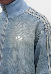 Άνδρας που φοράει ανοιχτό μπλε μπουφάν Adidas σε στυλ τζην με τρεις ριγωτές ραφές στον ώμο και φερμουάρ με ριγέ γιακά.