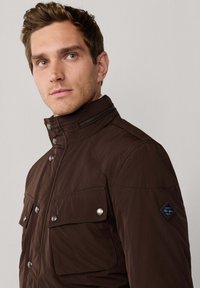 Mand iført en mørkebrun jakke med knaplukket klaplommer og en zip-høj krave, der vender en smule til venstre mod en simpel baggrund.