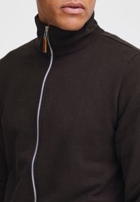 Mørkebrun zip-up genser med høy krage, med en sølvfarget glidelås og ribbede detaljer på halsen. Myk, teksturert stoff.