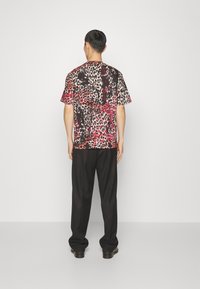 Man som står med ryggen mot kameran, klädd i en kortärmad skjorta med leopardmönster i röd och svart, svarta byxor och svarta skor.