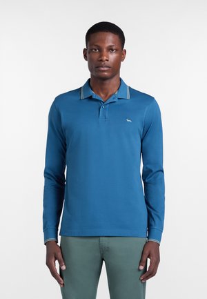 Muž v modrej polokošeli s dlhým rukávom s malým logom na hrudi a zelených nohaviciach, stojaci pred bielym pozadím.