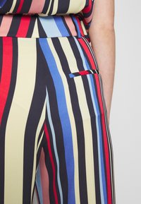 Pantalons rayés avec des lignes verticales en rouge, bleu, jaune et noir. Le matériau a une texture lisse ; comprend un détail de poche latérale.