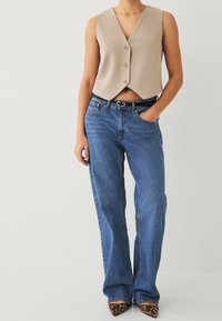 Beżowa kamizelka bez rękawów z dekoltem w serek i zapięciem na guziki, zestawiona z jasnoniebieskimi dżinsami o szerokich nogawkach oraz butami na wysokim obcasie w panterkę.