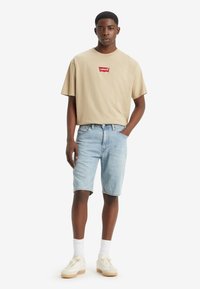 Beige cotton T-shirt with red Levi's logo, paired with light blue denim shorts and white high-top sneakers. Simple, casual design.