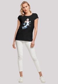 T-shirt noir avec un motif imprimé représentant un personnage aux cheveux clairs, vêtu d'une robe blanche et entouré de flocons de neige, associé à un pantalon blanc ajusté et des baskets.