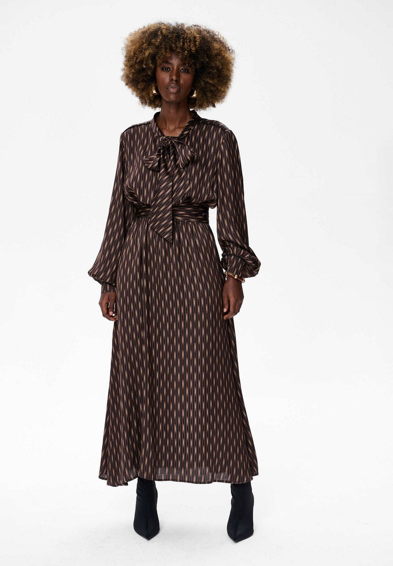 Maxi sukienka z ciemno-brązowego materiału w pionowe jasnobrązowe paski. Posiada sznurków w dekolcie oraz długie rękawy z dopasowanymi mankietami. Noszona z czarnymi botkami za kostkę.