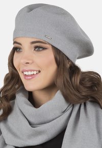 Berretto in maglia grigia con finitura testurizzata, abbinato a una sciarpa grigia coordinata. Presenta un piccolo dettaglio del logo in metallo sul berretto.