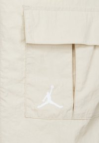Un tissu beige clair avec une grande poche plaquée et un logo Jumpman blanc brodé. La texture semble lisse avec de légères rides.