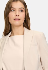 Beige op maat gemaakte blazer met gekantelde revers en een enkele borstzak, gecombineerd met een tops met hoge hals in lichtbeige. Glad stofoppervlak.