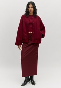 Bordeaux oversized jack met drukknoopjes, gedragen over een bijpassend aansluitend top en rok, gecombineerd met zwarte puntige laarzen. Glad textuur.