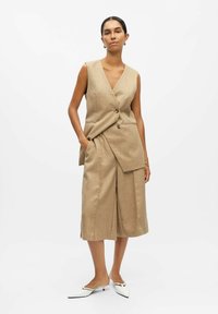 Gilet beige senza maniche con scollo a V, dotato di due bottoni e tasche frontali, abbinato a pantaloni corti a gamba larga, completato da décolleté bianchi a punta.