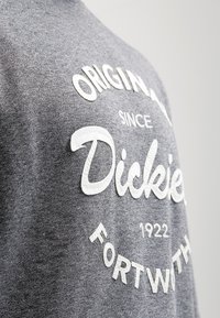 T-shirt grigio con testo ricamato bianco, che presenta "Dickies," "Original," "Dal 1922," e "Fort Worth," in design circolare.