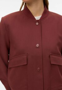 Veste bomber bordeaux en tissu lisse avec col côtelé, deux poches avant et boutons assortis. Design minimaliste.