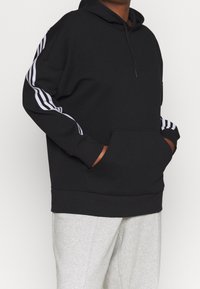 Personne portant un sweat à capuche noir avec des bandes blanches sur les manches et un pantalon gris, les mains dans la poche avant du sweat, sur un fond uni.