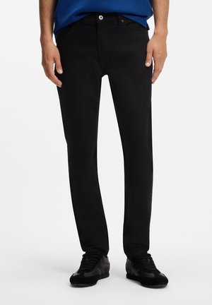 Uomo che indossa jeans neri aderenti, scarpe da ginnastica nere e una maglietta blu, in piedi davanti a uno sfondo bianco semplice.