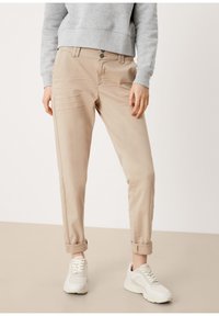 Osoba v béžových kalhotách s ohrnutými nohavicemi, bílých teniskách a světle šedé mikině, stojící proti jednoduchému neutrálnímu pozadí.