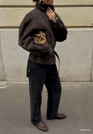 Femme portant une veste marron foncé oversized, un jean noir, des mocassins marron, tenant un sac à main beige avec une chaîne dorée, debout devant un mur beige.