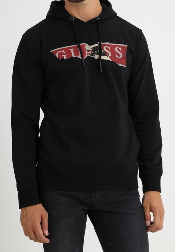 Homme portant un sweat à capuche noir avec un logo déchiré rouge et blanc "GUESS", associé à un jean foncé, debout sur un fond uni.
