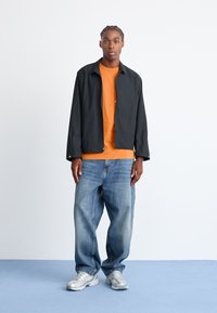 Sort letvægtsjakke over en orange t-shirt, parret med løse blå jeans og sølvfarvede sneakers, stående på en lys blå overflade.