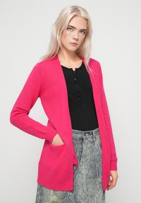 Cardigan fucsia con apertura frontale, maniche lunghe, polsini a coste e due tasche frontali, abbinato a una maglia nera e una gonna a fantasia.