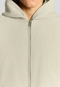 Ljusbeige zip-up hoodie med en mjuk textur, utrustad med ett fodrat huva och silverfärgad dragkedja med varumärkesdetalj vid halslinningen.