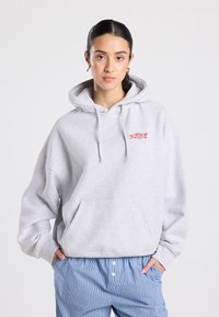 Sweat à capuche oversize gris clair en mélange de coton avec une poche frontale et une capuche ajustable à cordon. Présente un petit motif rouge sur la poitrine.