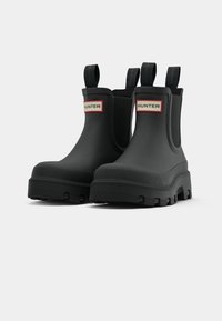 Botas de goma negras hasta el tobillo con un acabado texturizado, que presentan tiradores y una etiqueta roja y blanca "HUNTER" en el lateral. Suelas gruesas y con patrones.
