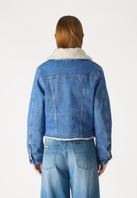 Niebieska dżinsowa kurtka z białym kołnierzem i mankietami z baranka, z krojem krótkim i widocznymi detalami szycia. Noszona z niebieskimi dżinsami.
