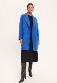 Niebieski płaszcz z wełny syntetycznej, zapinany na dwa guziki, z klapami, bocznymi kieszeniami i luźnym krojem, zestawiony z czarną sukienką i wysokimi butami.