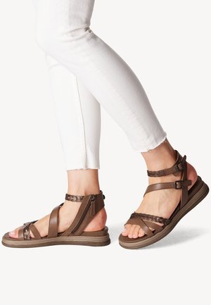 Fødder iført brune læder sandaler med stropper, sidelommer med lynlåse og spænder, kombineret med hvide cropped bukser mod en ensfarvet baggrund.