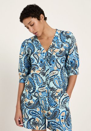 Woman wearing a blue paisley-patterned V-neck blouse with three-quarter sleeves and matching shorts, with one hand in pocket.