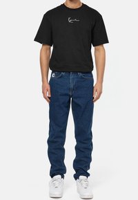 Hombre que lleva una camiseta negra de manga corta con logotipo, jeans azules con parche de logotipo y zapatillas blancas, de pie contra un fondo blanco.