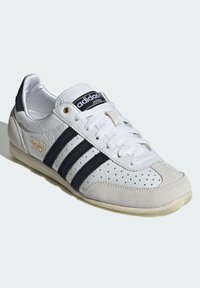 Sneakers din piele albă cu accente din suede negru și trei dungi negre. Prezintă vârful perforat, talpa din cauciuc texturat și branding auriu.